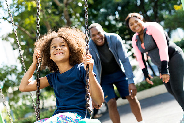 A young girl plays on a swing while her parents smile in the background.